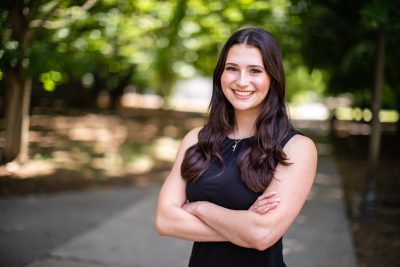 Portrait of Jess Gannon, graduate student in biomedical engineering. She stands in front of blurred-out trees on campus, in a black sleeveless dress.
